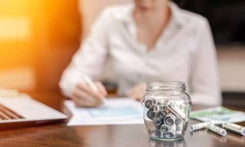 íróasztalnál fehér ingben egy nő kimutatásokat néz, előtérben befőttesüvegben pénzkötegek gyűlnek