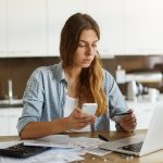 fiatal nő laptoppal és mobillal online ügyeket intéz a konyhában