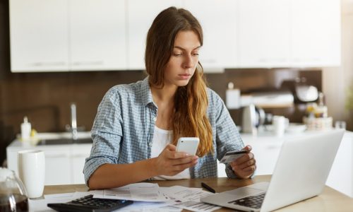 fiatal nő laptoppal és mobillal online ügyeket intéz a konyhában