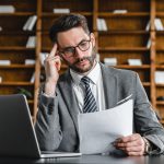 öltönyös vállalkozó irodai környezetben papírokat olvas laptop előtt